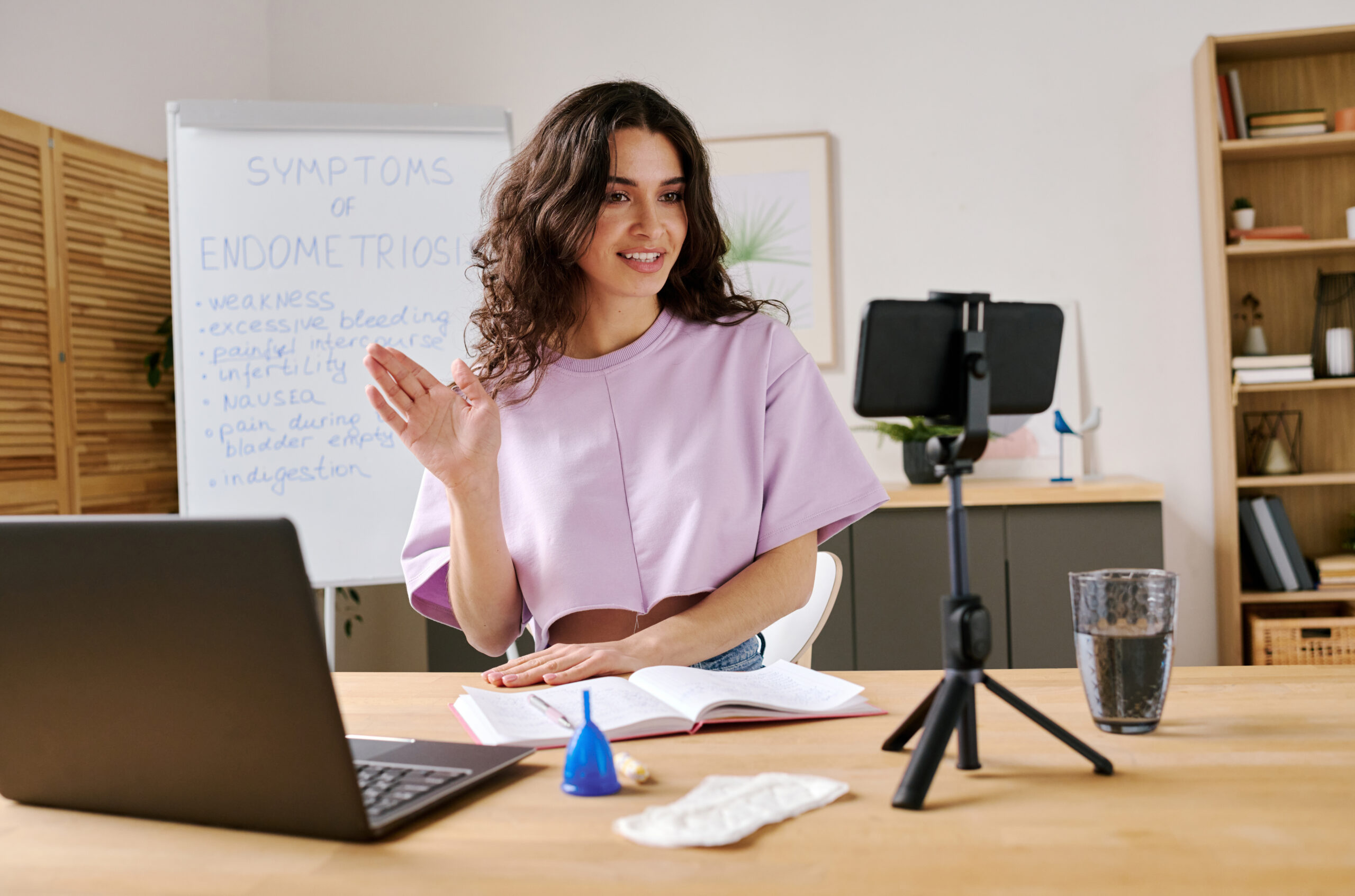 Young woman sitting at desk in front of smartphone on tripod starting live stream waving hand to her followers