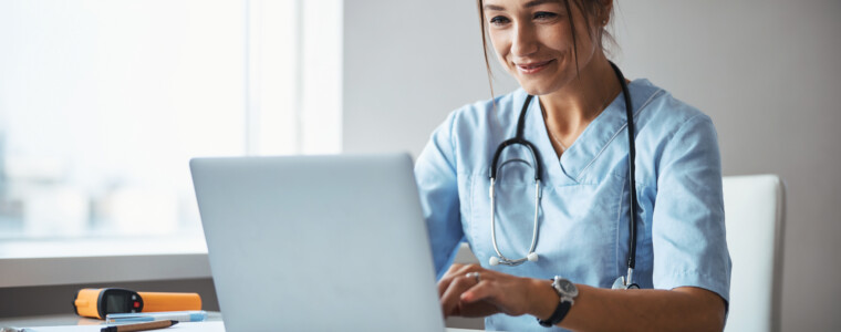 Beautiful young woman physician sitting at the table and smiling while working on modern laptop