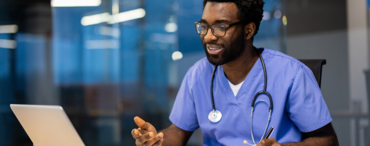 A black male doctor in scrubs and stethoscope smiles while taking notes during a telehealth appointment in his modern office.