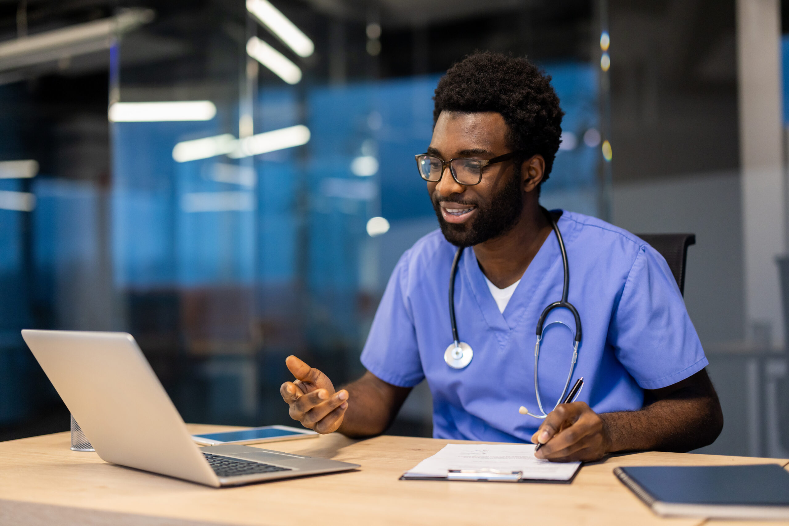 A black male doctor in scrubs and stethoscope smiles while taking notes during a telehealth appointment in his modern office.