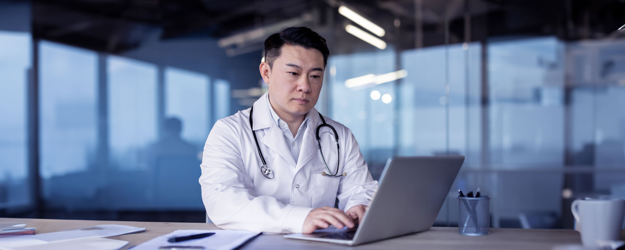 Serious young Asian male doctor in white coat working intently at laptop.