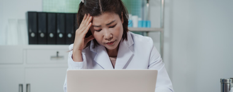 Stressed female doctor in white coat working on laptop at clinic desk, holding head with hand, paperwork and stethoscope nearby, healthcare burnout concept