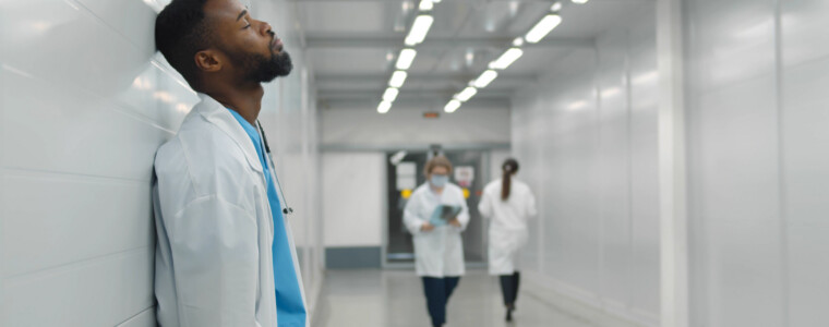 Tired male african doctor standing in hospital corridor. Portrait of exhausted and stressed afro-american medical worker leaning on wall and resting in clinic hallway
